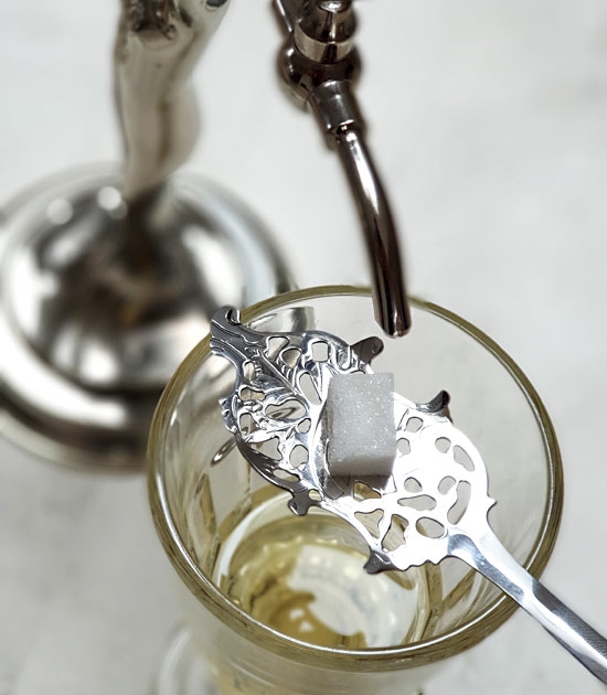 Absinthe wormwood spoon sitting on absinthe glass, water dripping metal absinthe fountain.