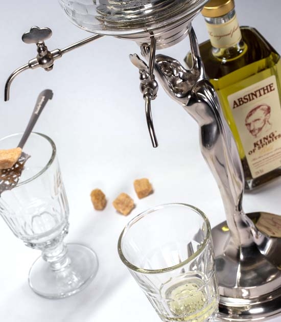 Metal absinthe fountain with bottle of premium absinthe, glass, absinthe spoon, and sugar cubes.